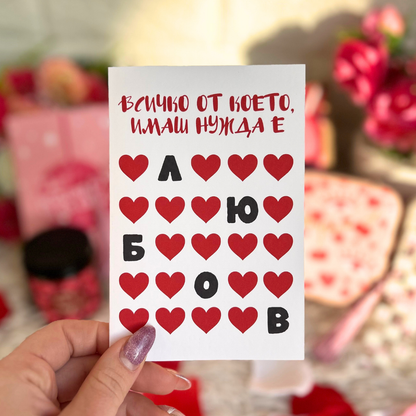 Ръка държи картичка с надпис „Всичко от което имаш нужда е любов“ с червени сърца – романтичен детайл към подарък за Свети Валентин